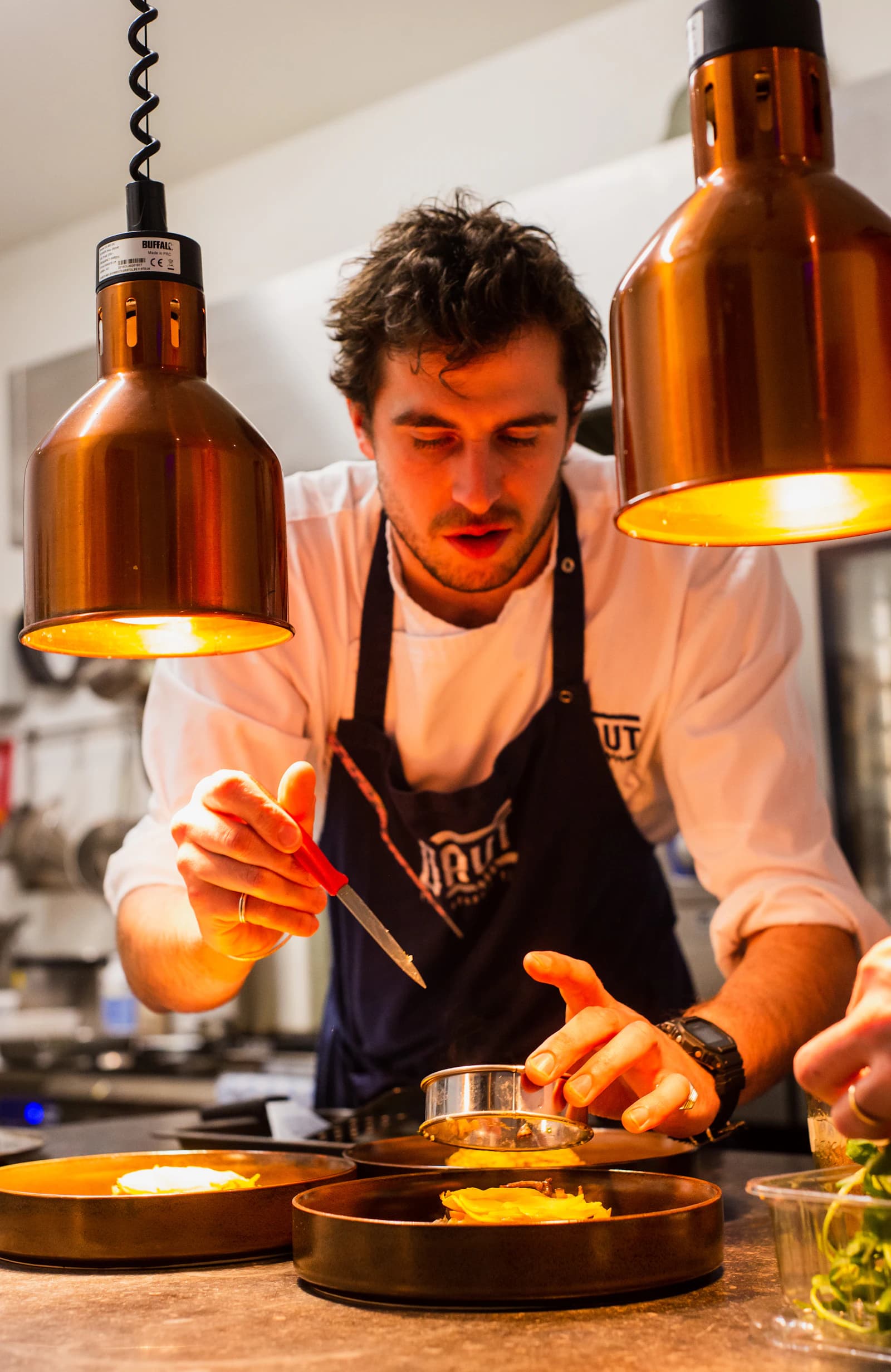 A chef plating a dish with tweezers under copper pass lamps.