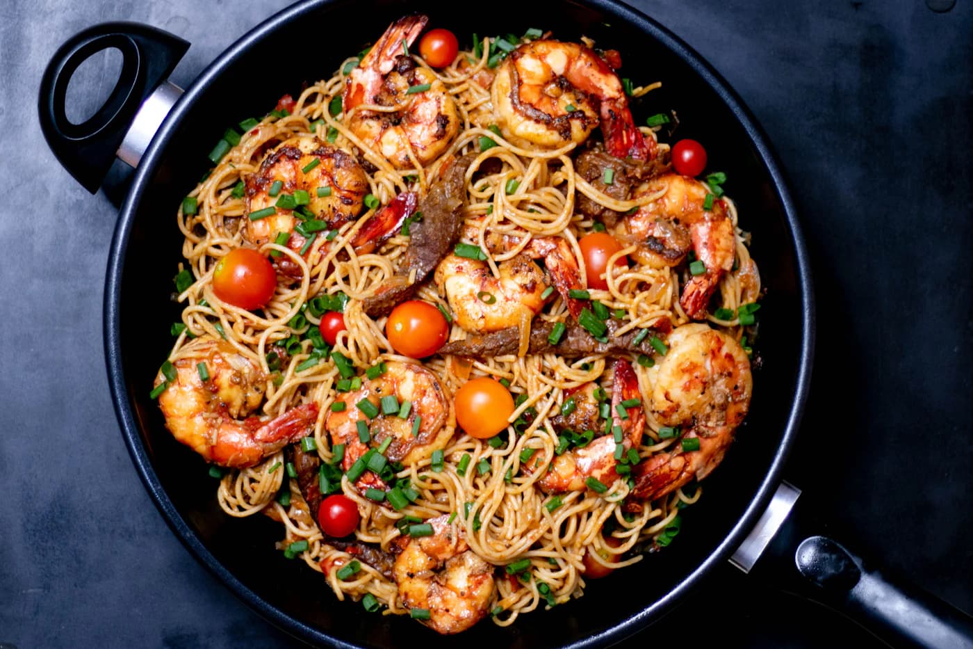 Linguine tossed with prawns and cherry tomatoes in a black pan.