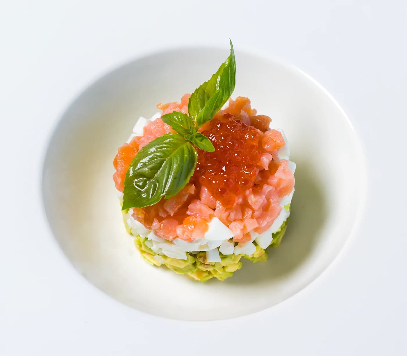 Trout tartare crowned with orange roe and a basil leaf in a white bowl.
