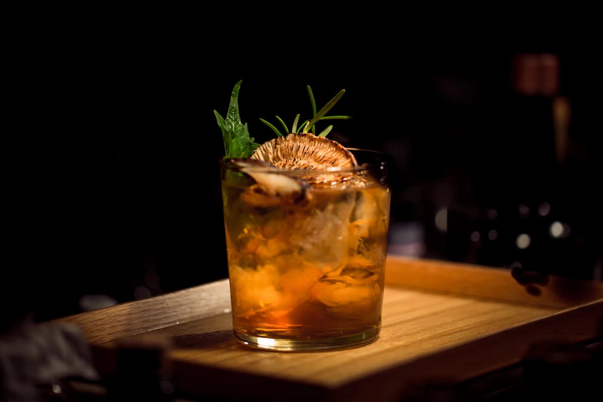 An amber cocktail garnished with a rosemary sprig on a wooden bar.