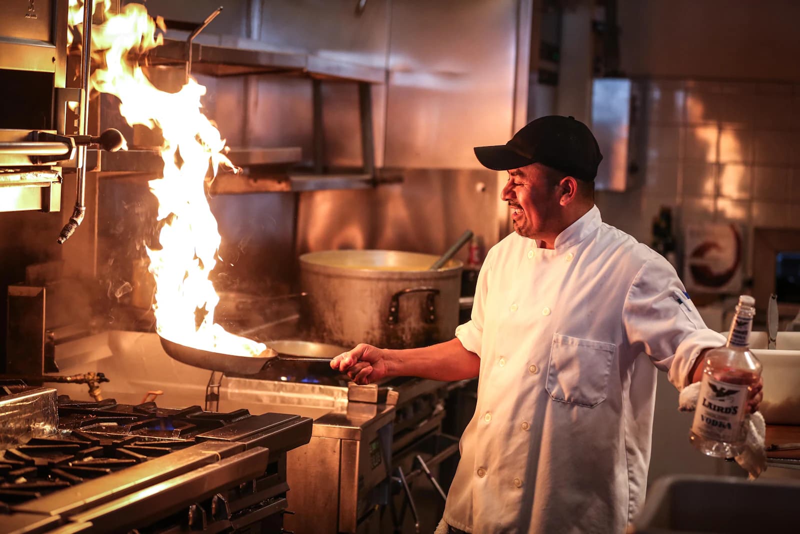A cook flames a pan over a high burner in a busy kitchen.
